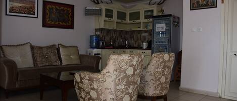 A living room with a sofa, two armchairs, a coffee table, and a kitchen area with cabinets and a refrigerator.