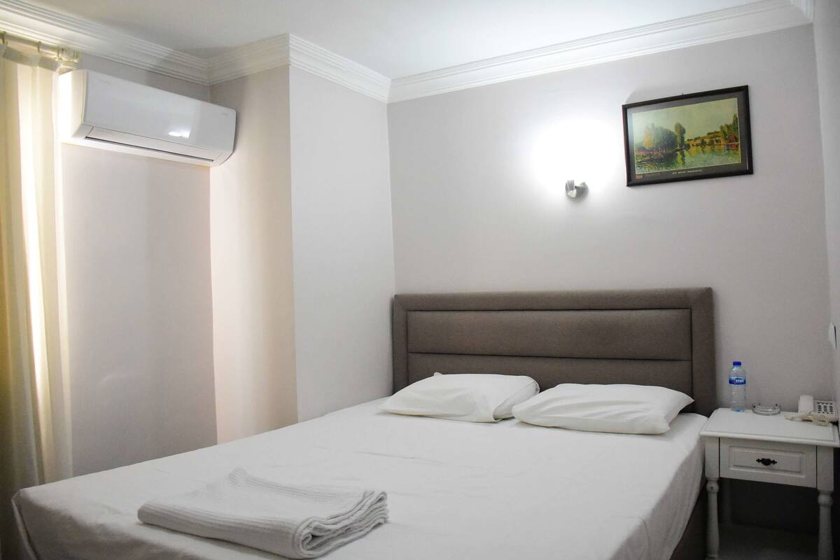 Hotellihuone, jossa on sänky, yöpöytä, vesipullo ja taulu seinällä.