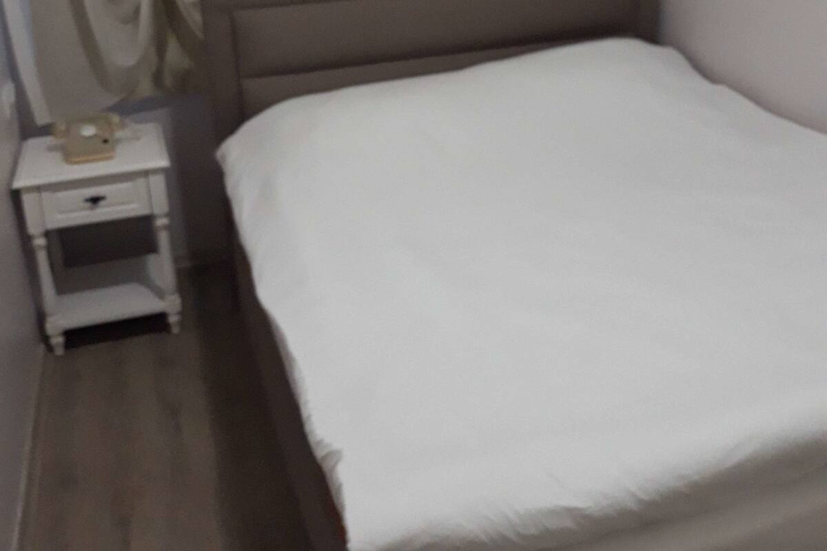 Siististi pedattu sänky hotellihuoneessa, jossa on puinen pääty, pieni yöpöytä lampun kanssa ja verho, joka on osittain vedetty sivuun.