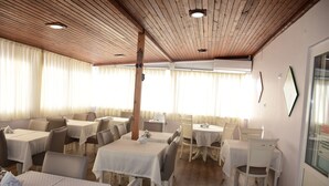 Zona de dinar amb diverses taules i cadires, sostre de fusta i grans finestres amb cortines.