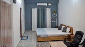 Une chambre à coucher comprenant un lit, une chaise, une table, un ventilateur de plafond, un climatiseur, une horloge et une fenêtre avec des rideaux.