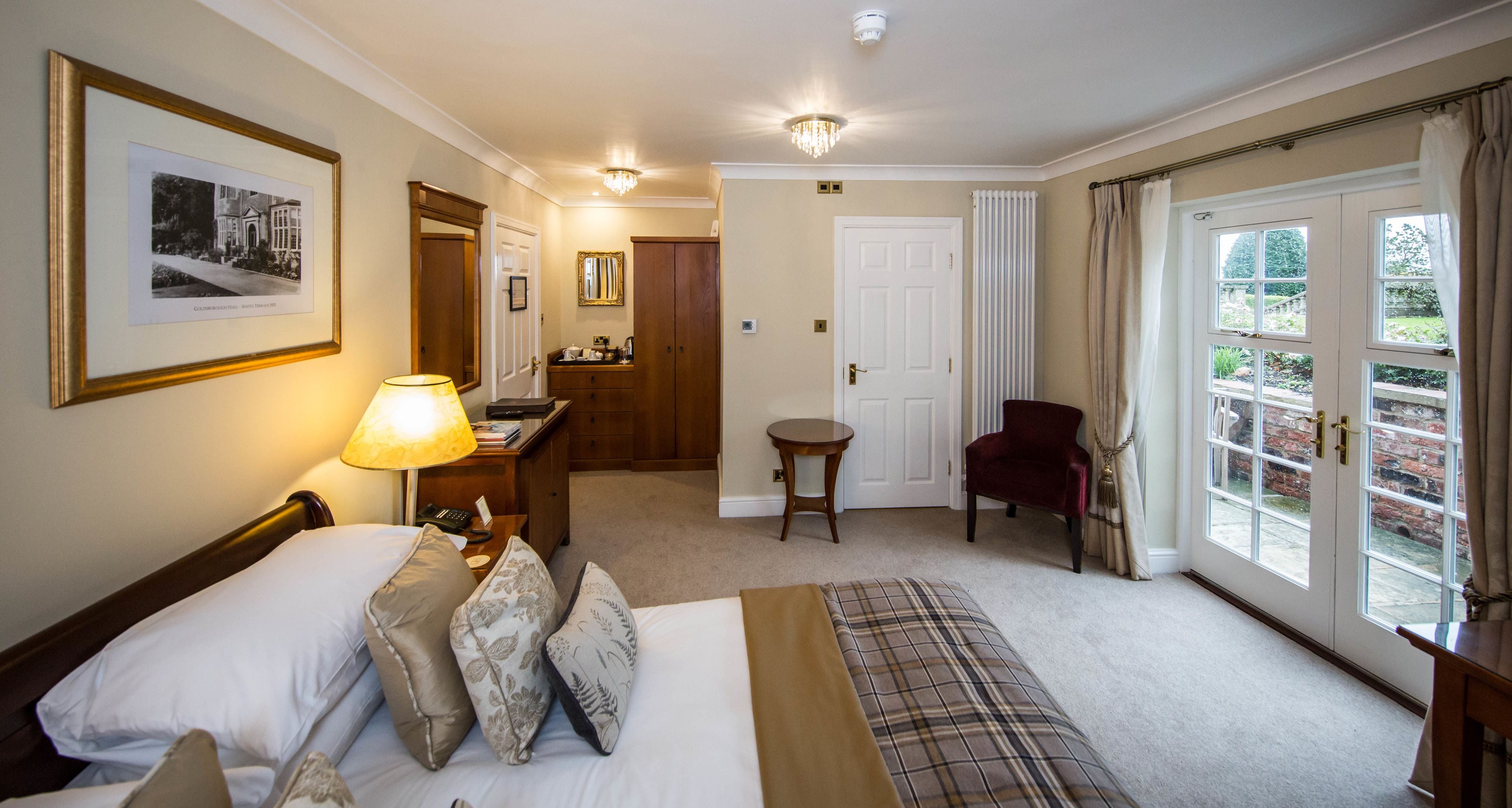 A hotel room with a bed, bedside table, lamp, and a view of the garden through the door.