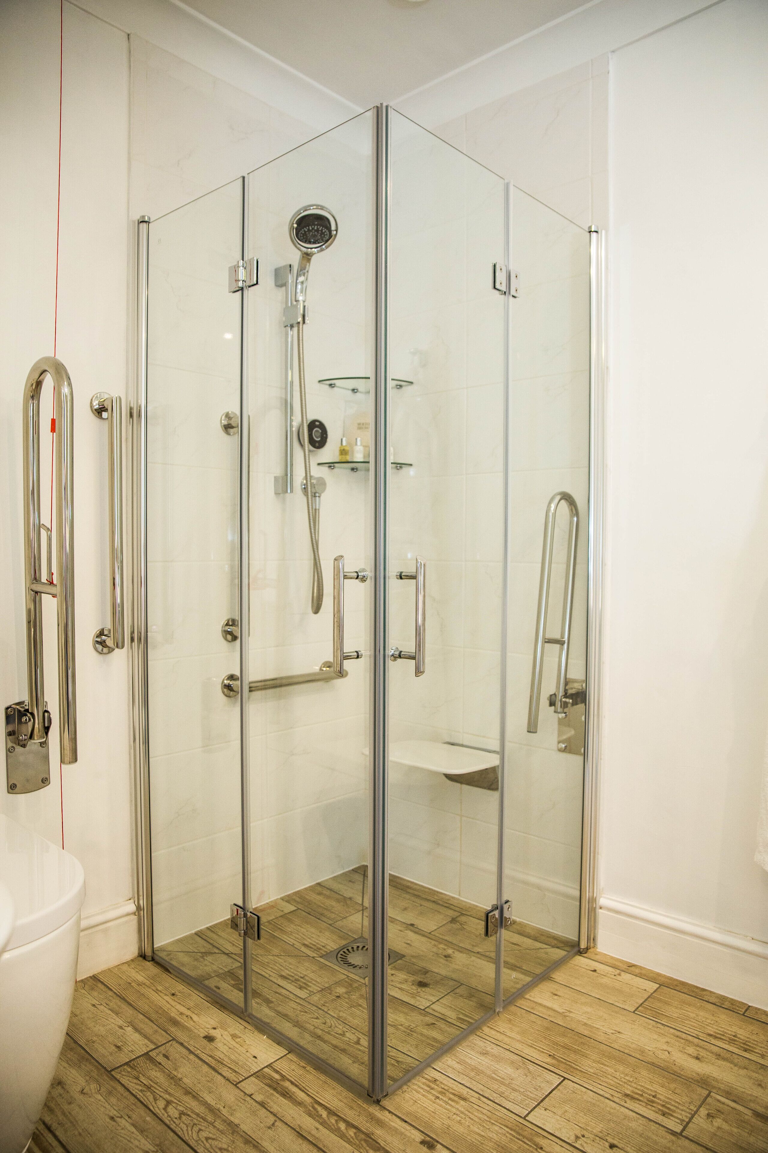 A glass-enclosed shower with a handheld showerhead, a handheld faucet, and a glass shelf.