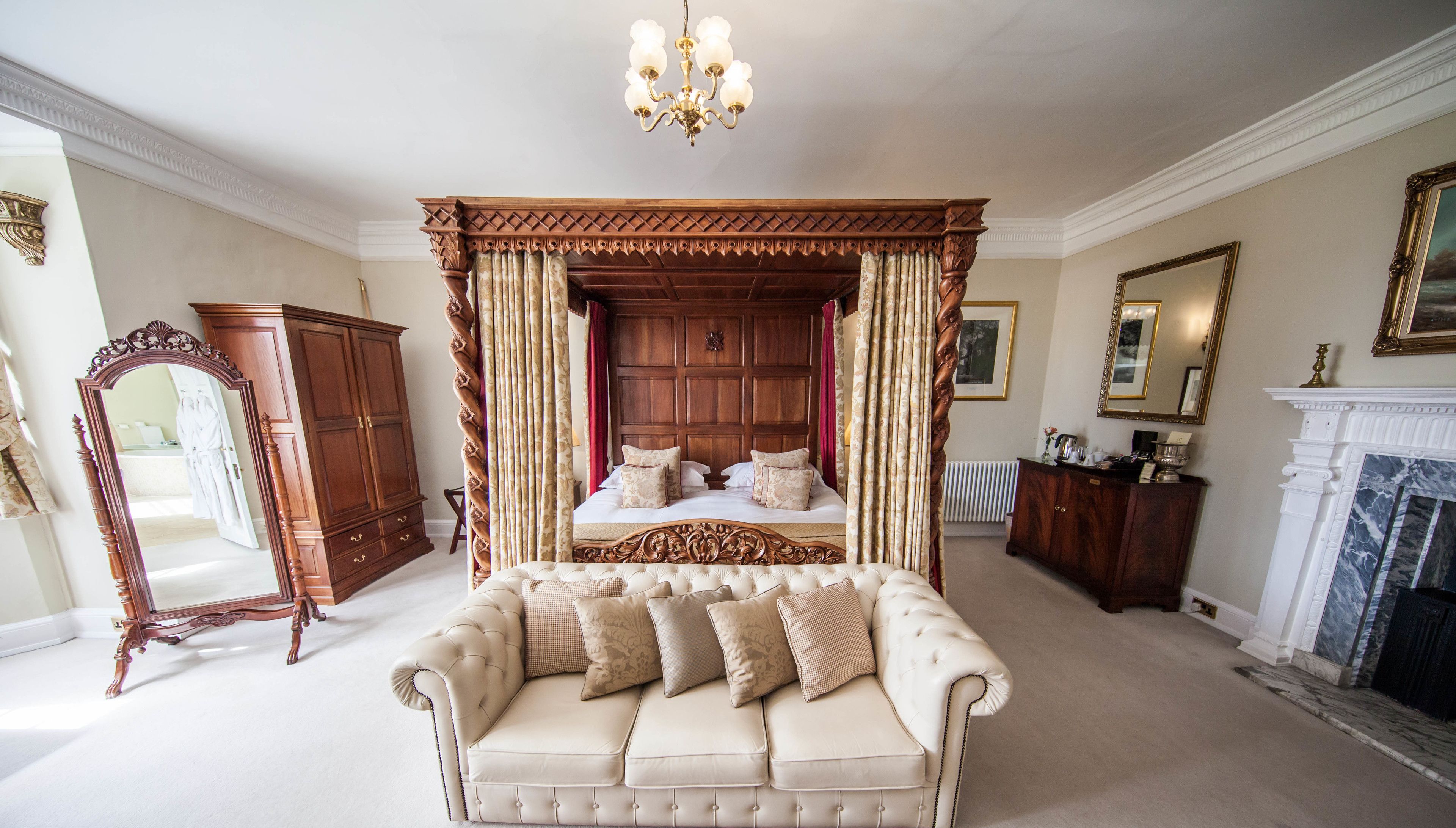 A bedroom with a four-poster bed, a chaise lounge, a mirror, a fireplace, and a large wooden wardrobe.
