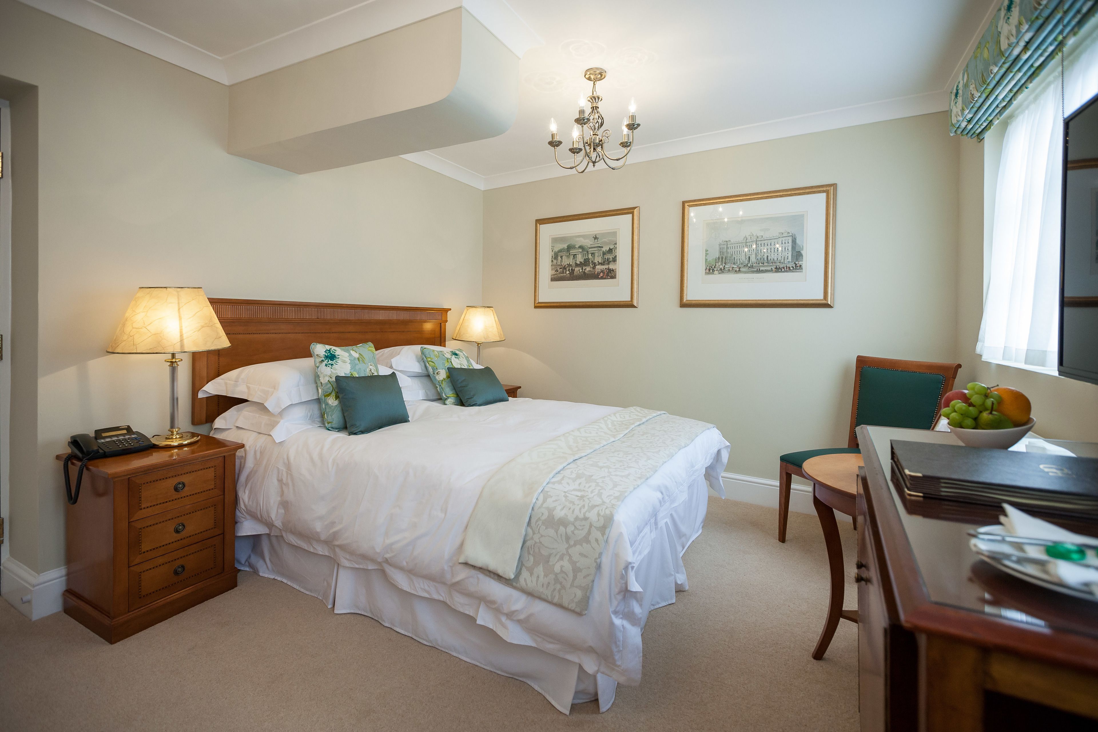 A neatly arranged bedroom with a large bed, bedside lamps, a wooden nightstand, a desk with a chair, and two framed pictures on the wall.