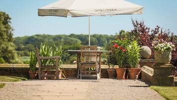 A wooden bench and table under a Nyetimber umbrella, surrounded by potted plants and a gravel path.