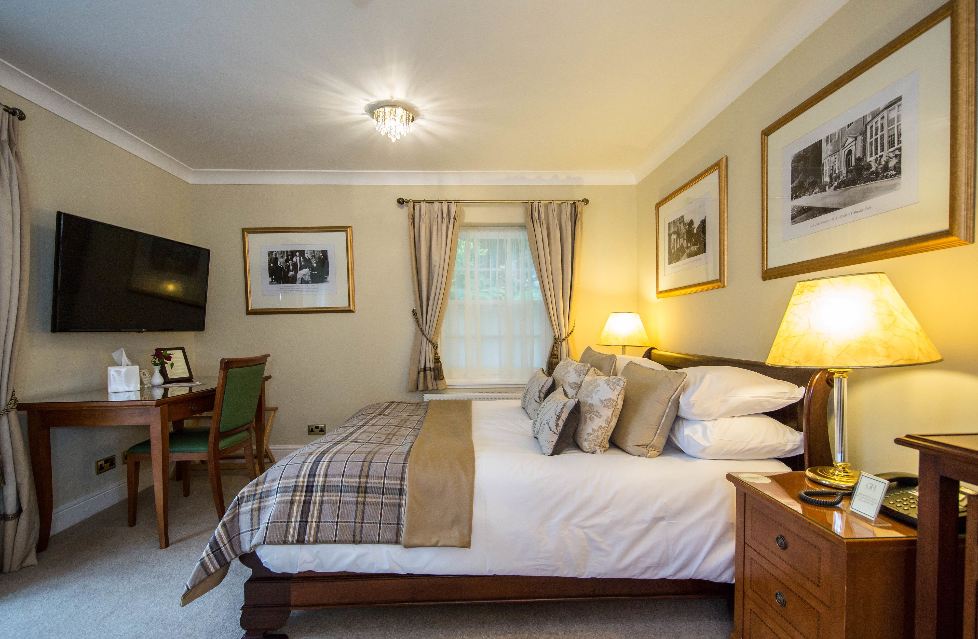 A hotel room with a bed, bedside table, lamp, chair, desk, TV, and framed pictures on the wall.