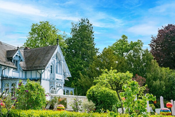 A large, white, Tudor-style building with a steeply pitched roof and multiple chimneys, surrounded by lush greenery and trees.
