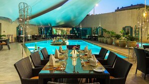 Zona de piscina al aire libre con mesas y sillas para comer, rodeada de plantas en macetas y un paraguas grande.