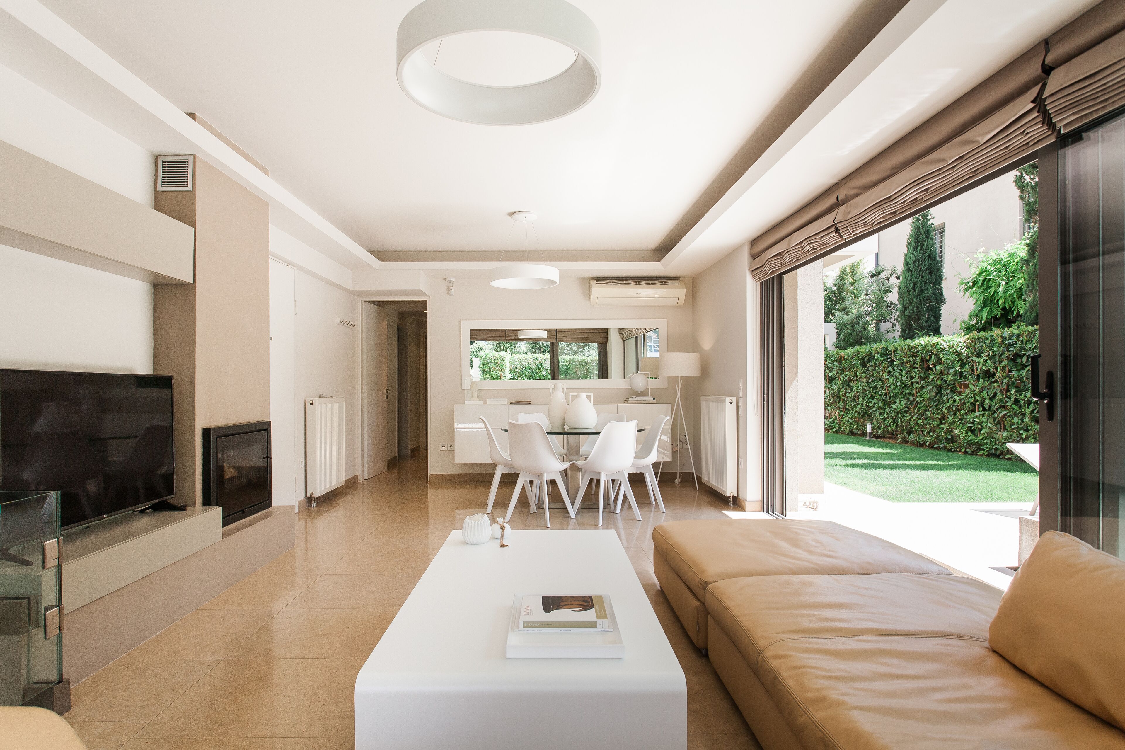 A modern living room with a flat-screen TV, a white coffee table, and a beige sofa. There is a dining area with a white table and chairs, and a kitchen visible in the background.