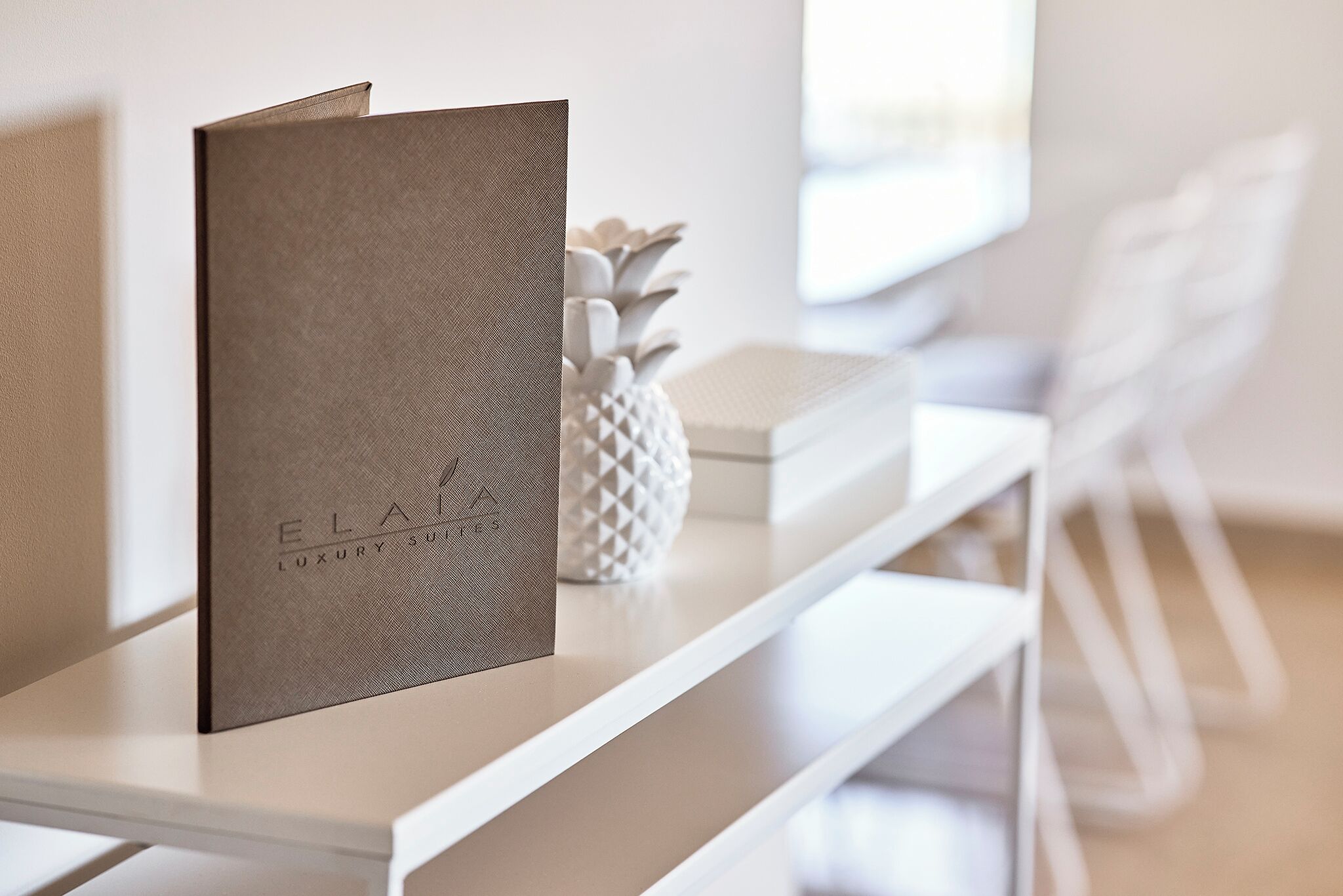 Ein moderner, minimalistischer Raum mit einem weißen Konsolentisch, einer dekorativen Ananas und einem gerahmten Schriftzug 'ELLA LUXURY SUITES'.