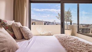 A bedroom with a large bed, a fluffy rug, a chair, and a view of the city through a large window. - Crystal Green Signature Apartments Glyfada (Glyfada)