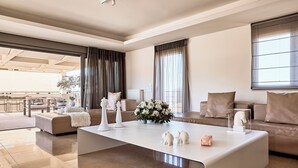 A modern living room with a central white coffee table, a sofa, and a dining area visible through a large window with curtains. - Crystal Green Signature Apartments Glyfada (Glyfada)