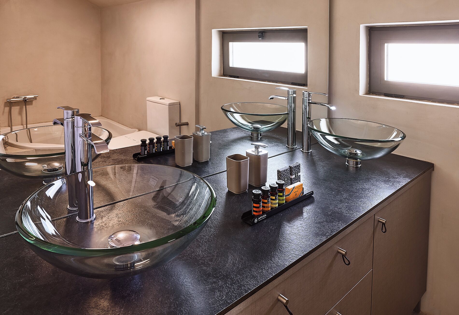 A modern bathroom with a glass vessel sink, a handheld showerhead, and a built-in toilet. There are two windows providing natural light.