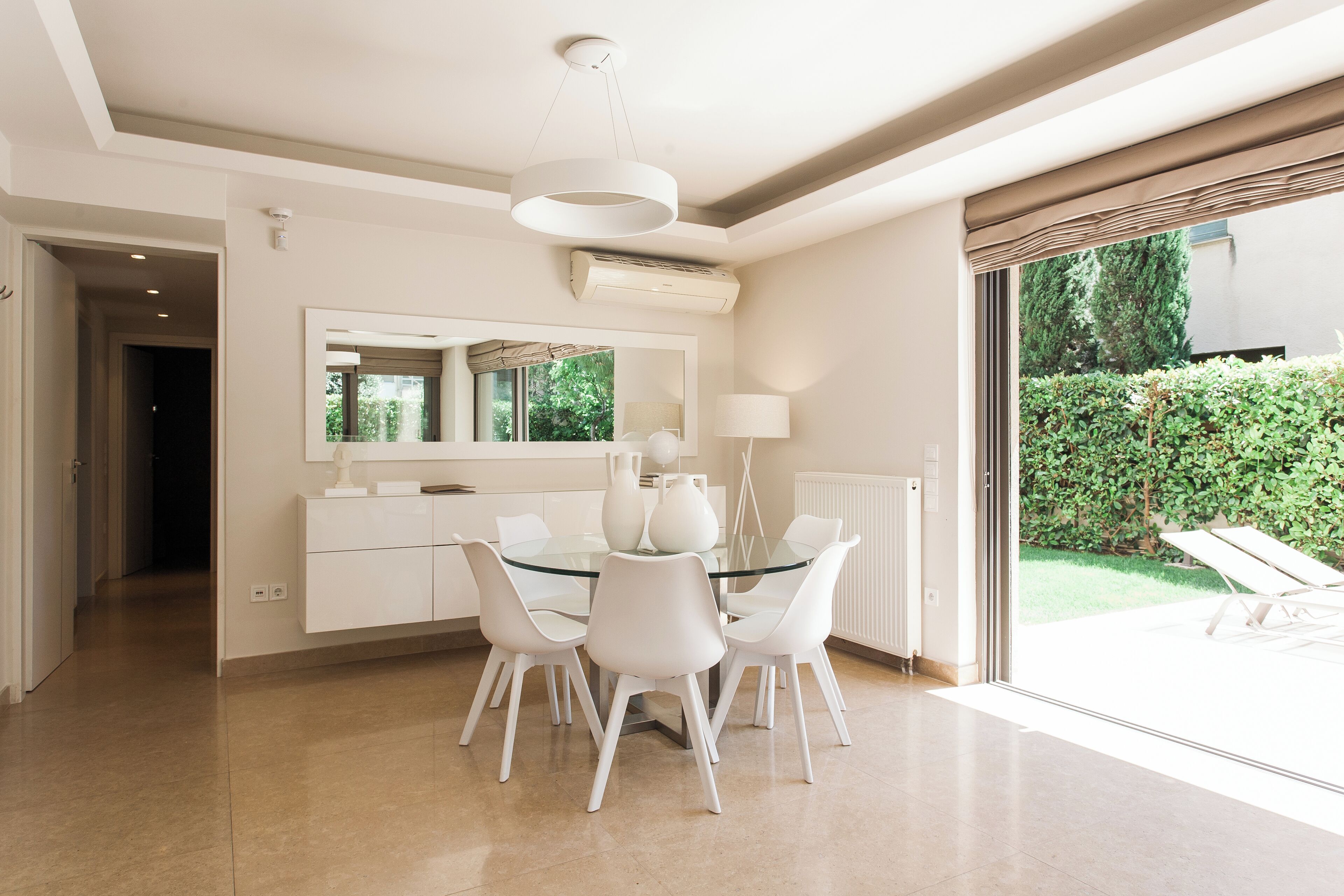 A modern dining area with a glass table and white chairs, a large mirror above the table, and a view of a garden through a sliding glass door.