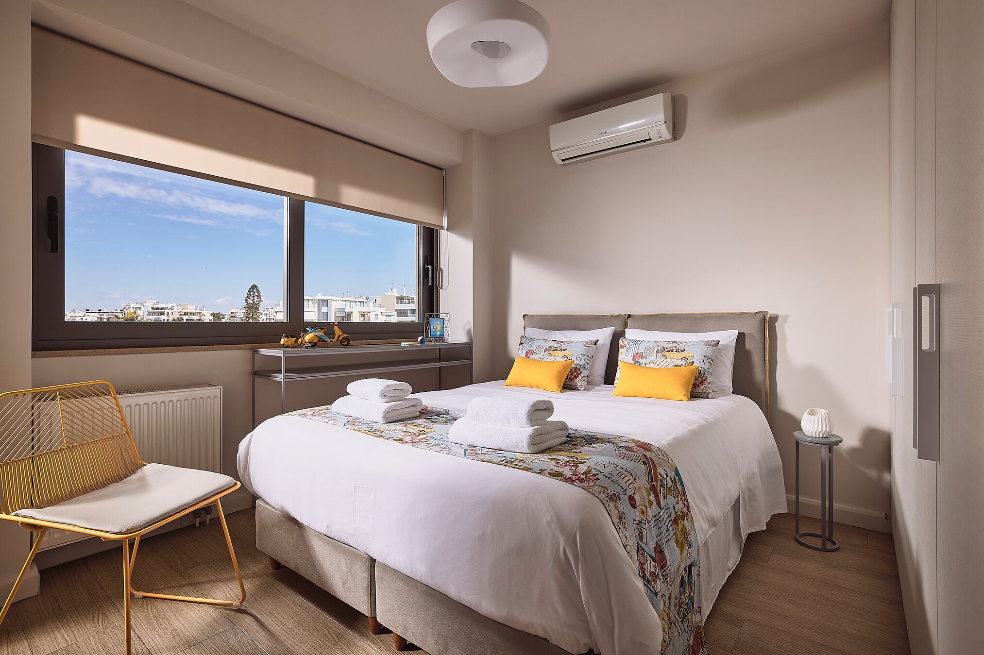 A modern hotel room with a large bed, a chair, a small table, and a view of the city through large windows.