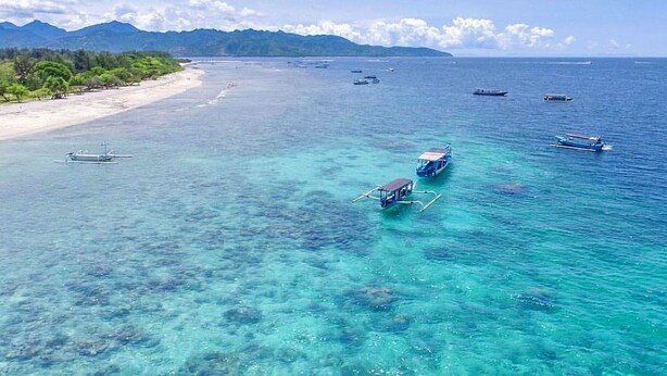 Una playa con agua turquesa cristalina, embarcaciones y una cadena montañosa distante.
