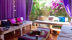 Una habitación acogedora con sofá, mesa y banco. Cortinas moradas y vistas a un jardín con flores rosadas.