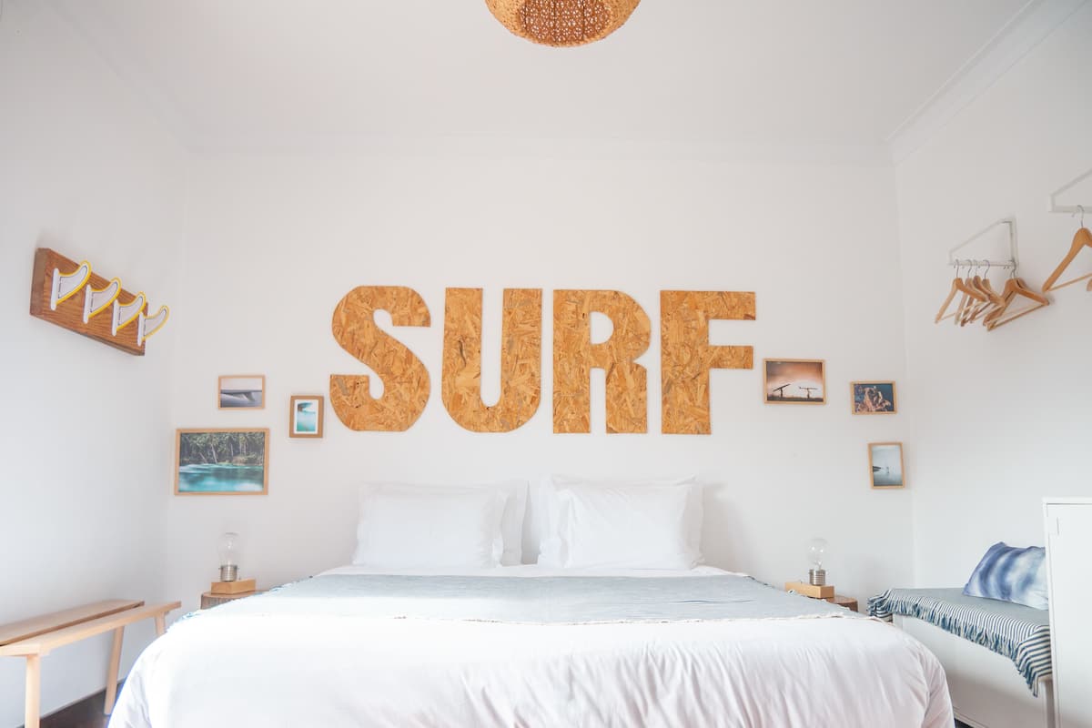 A bedroom with a large corkboard wall art spelling "SURF", a bed with white bedding, a bedside table, and a hanging light fixture.