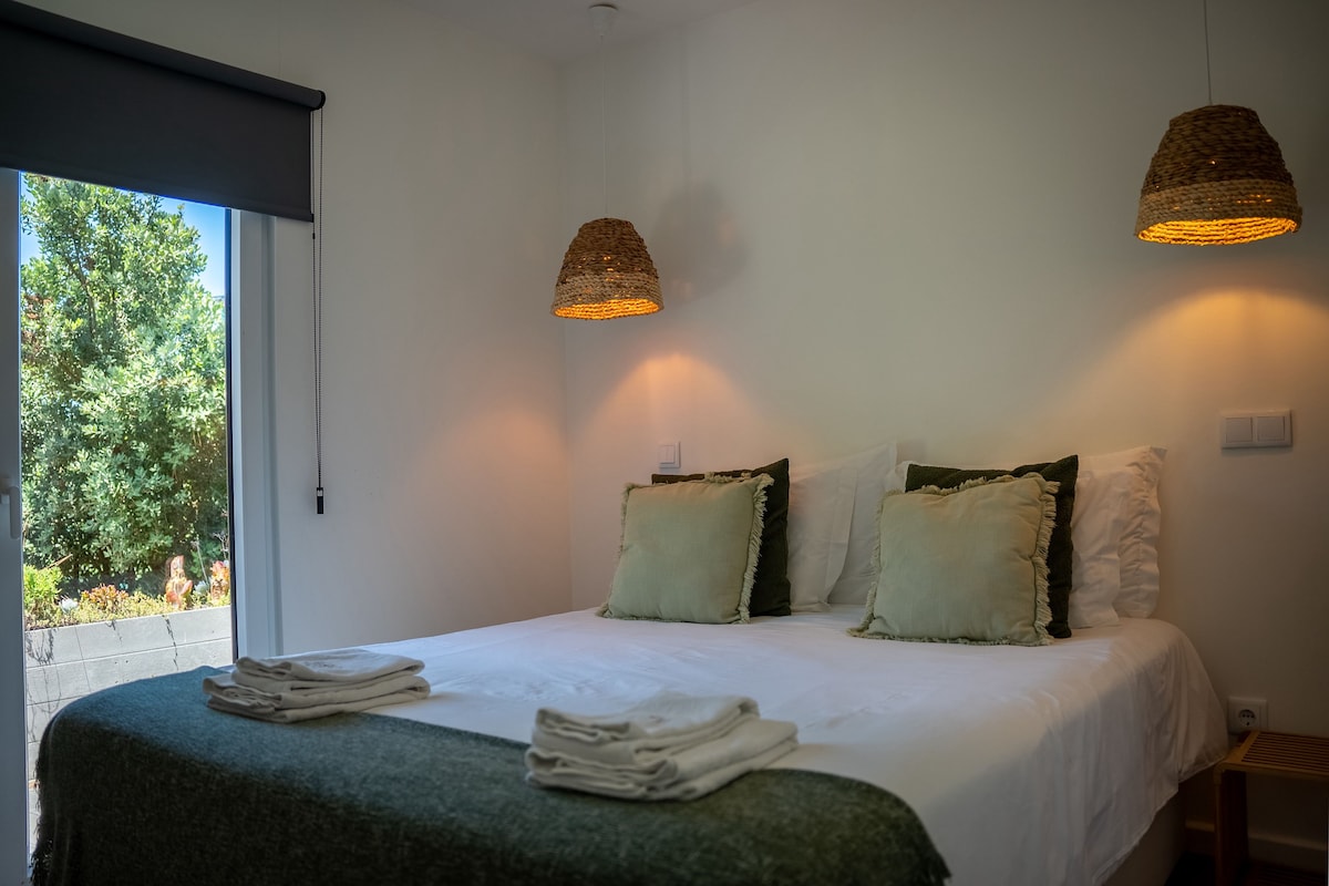 A bedroom with a bed, two wall-mounted lamps, a window, and a view of trees.