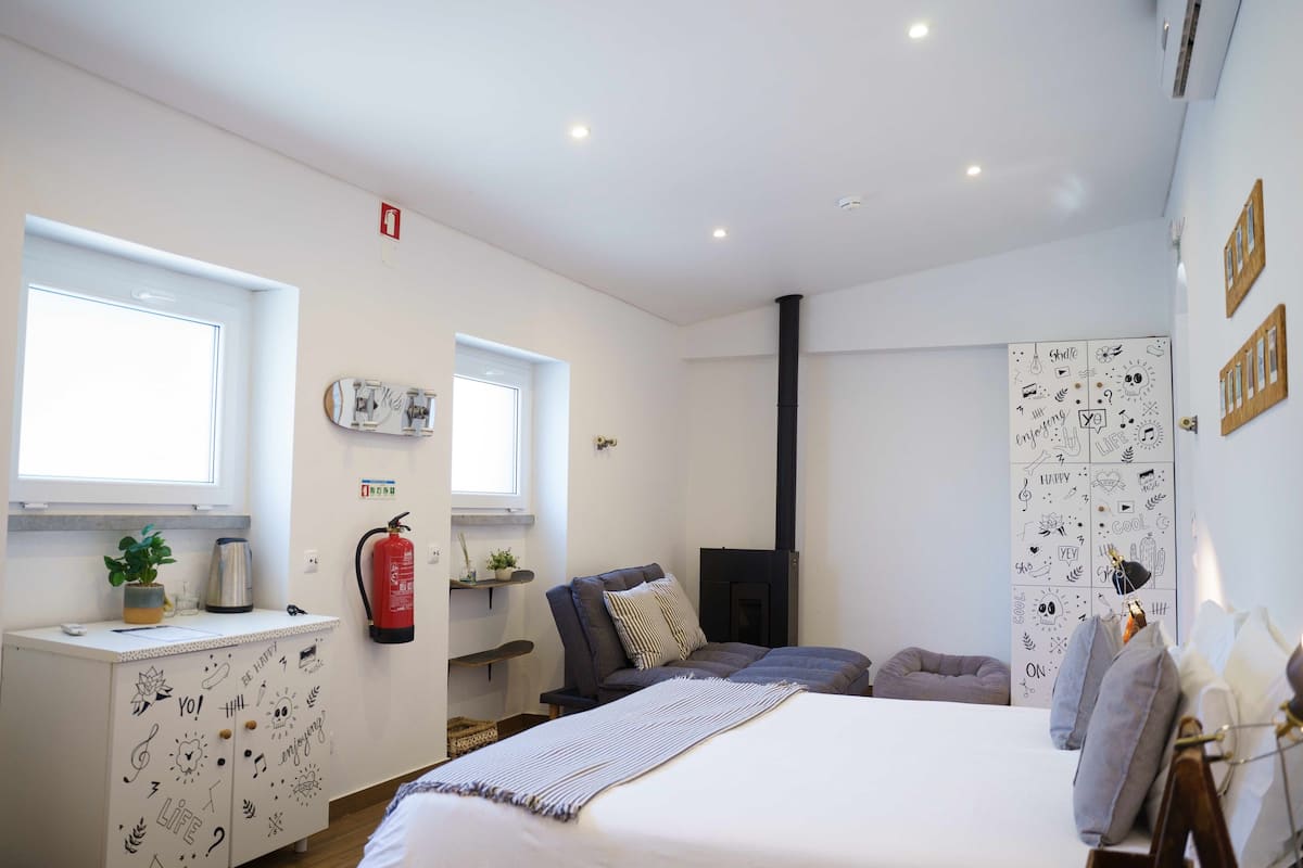A bedroom with a bed, a chair, a fireplace, a shelf with plants, and a window.