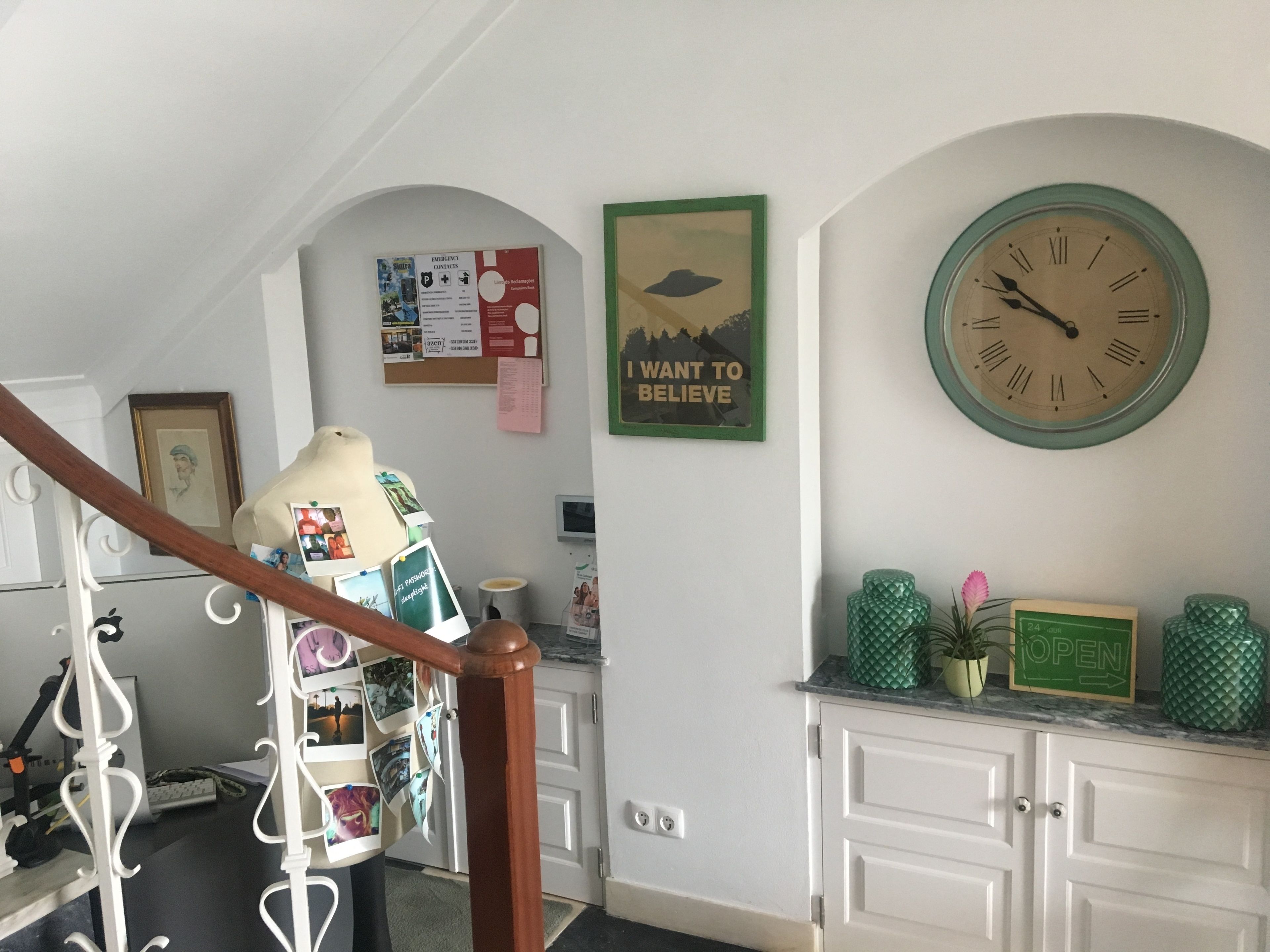 Una escalera con barandilla de madera, un tablón de anuncios con varios avisos, un cartel enmarcado y un reloj de pared.