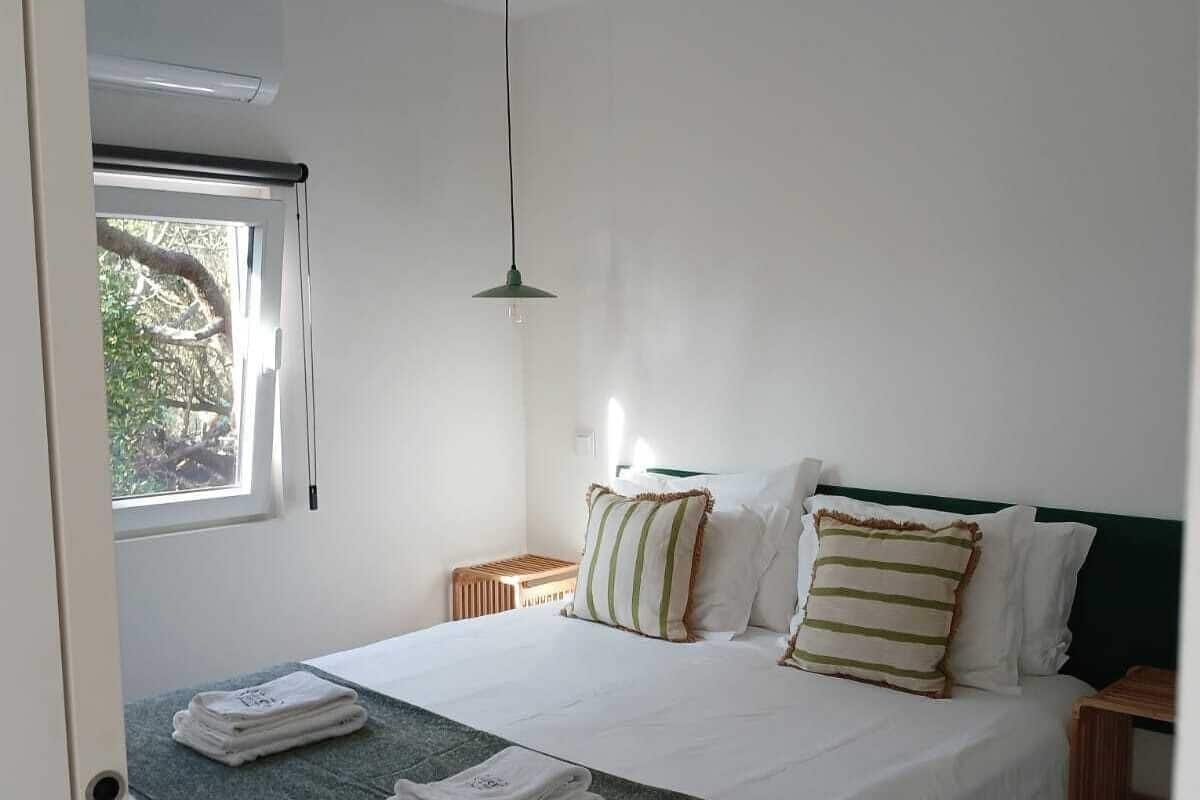 A neatly made bed with white linens and striped pillows, a green throw blanket, and a window with a view of trees.