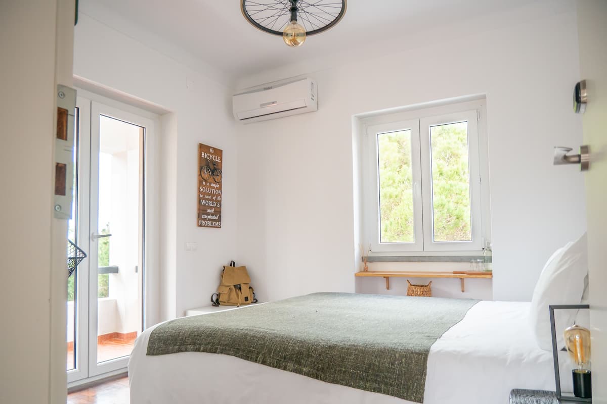 A bedroom with a bed, a window, a wall-mounted air conditioner, and a door leading to an outdoor area.