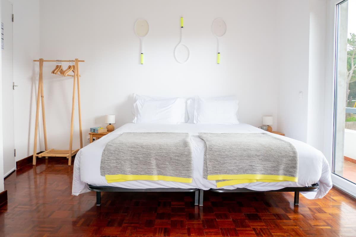 A double bed with a grey and yellow blanket, a wooden floor, a wooden hanger rack, and a window with a view.