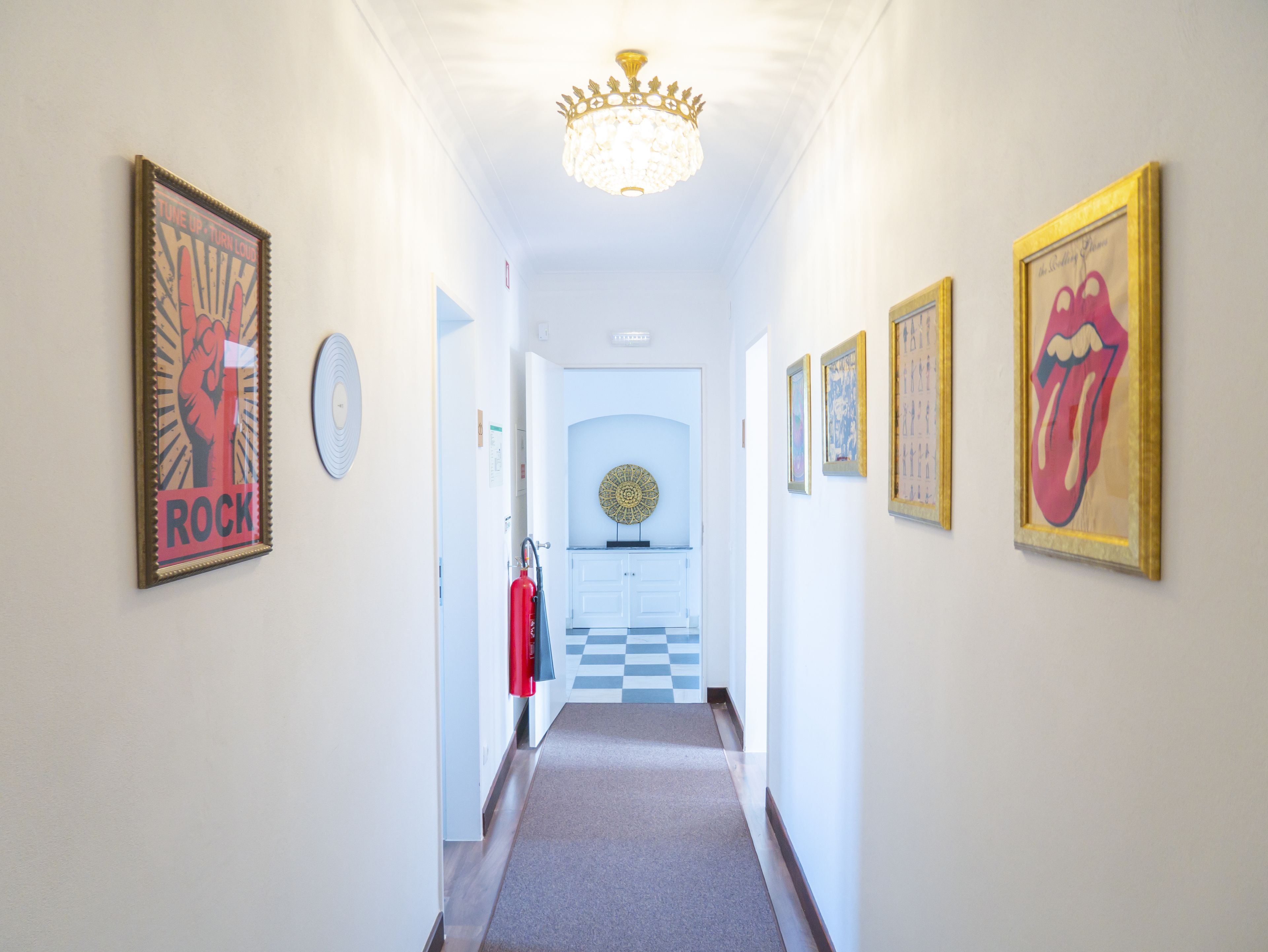 A hallway with framed posters, a chandelier, and a checkered floor leading to a room.