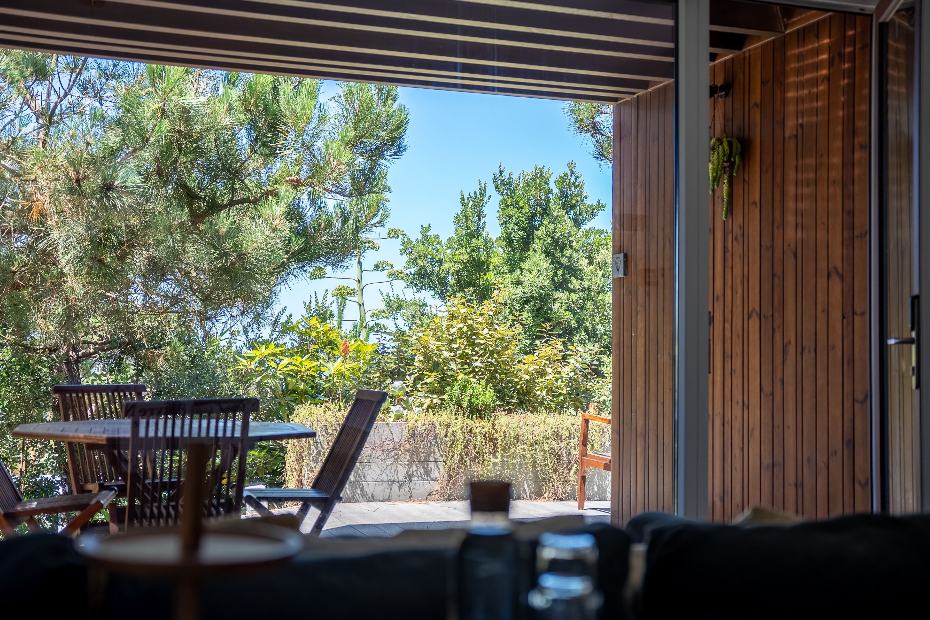Ein überdachter Außenbereich mit Tisch und Stühlen, mit Blick auf einen Garten mit verschiedenen Bäumen und Pflanzen.