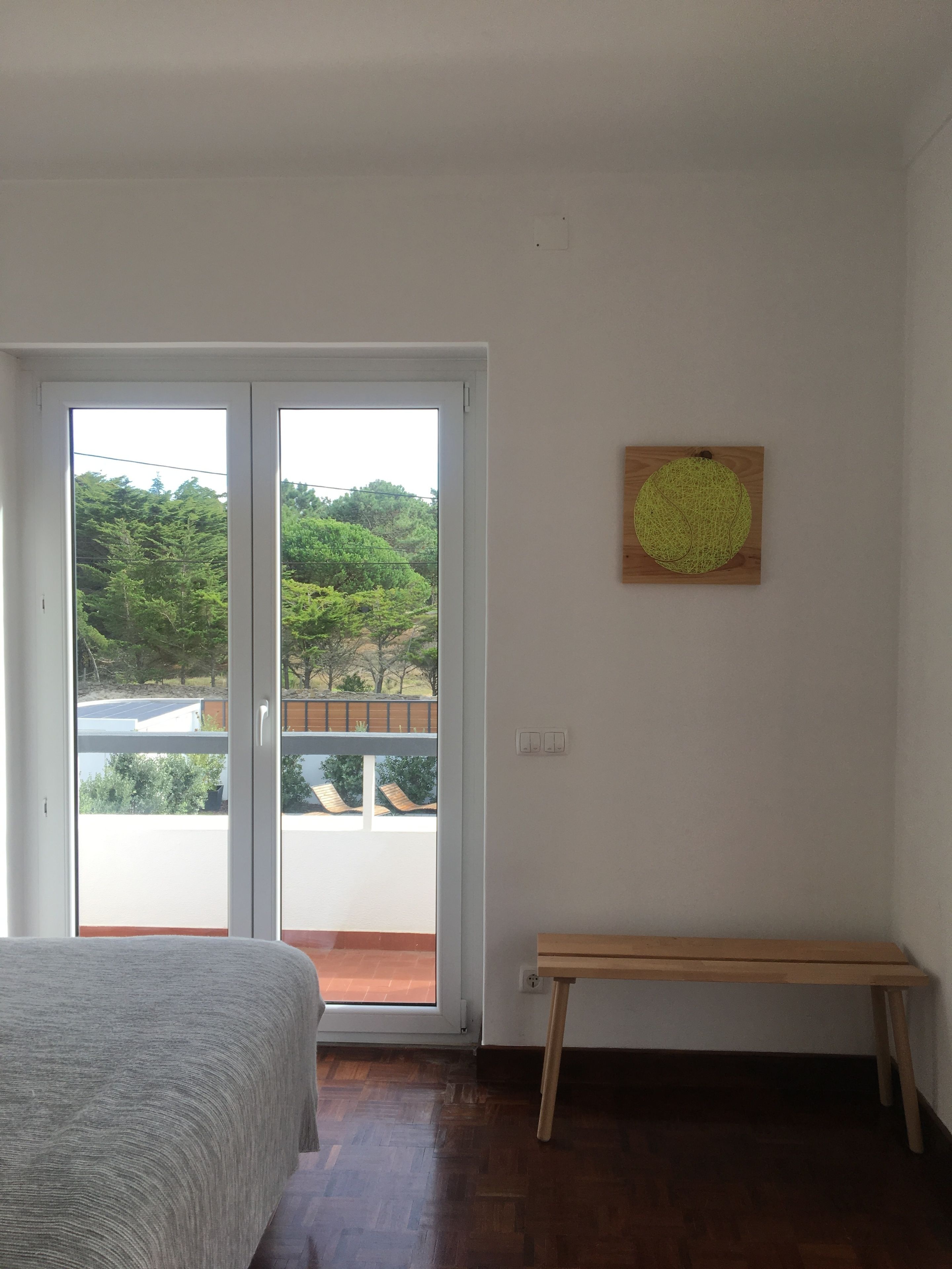 Un dormitorio con cama, un banco, una puerta corrediza de vidrio que da a un balcón y una obra de arte colgada en la pared.