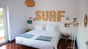 Quarto com uma cama grande, um letreiro fixado na parede com a palavra 'SURF', piso de madeira e porta com espelho.