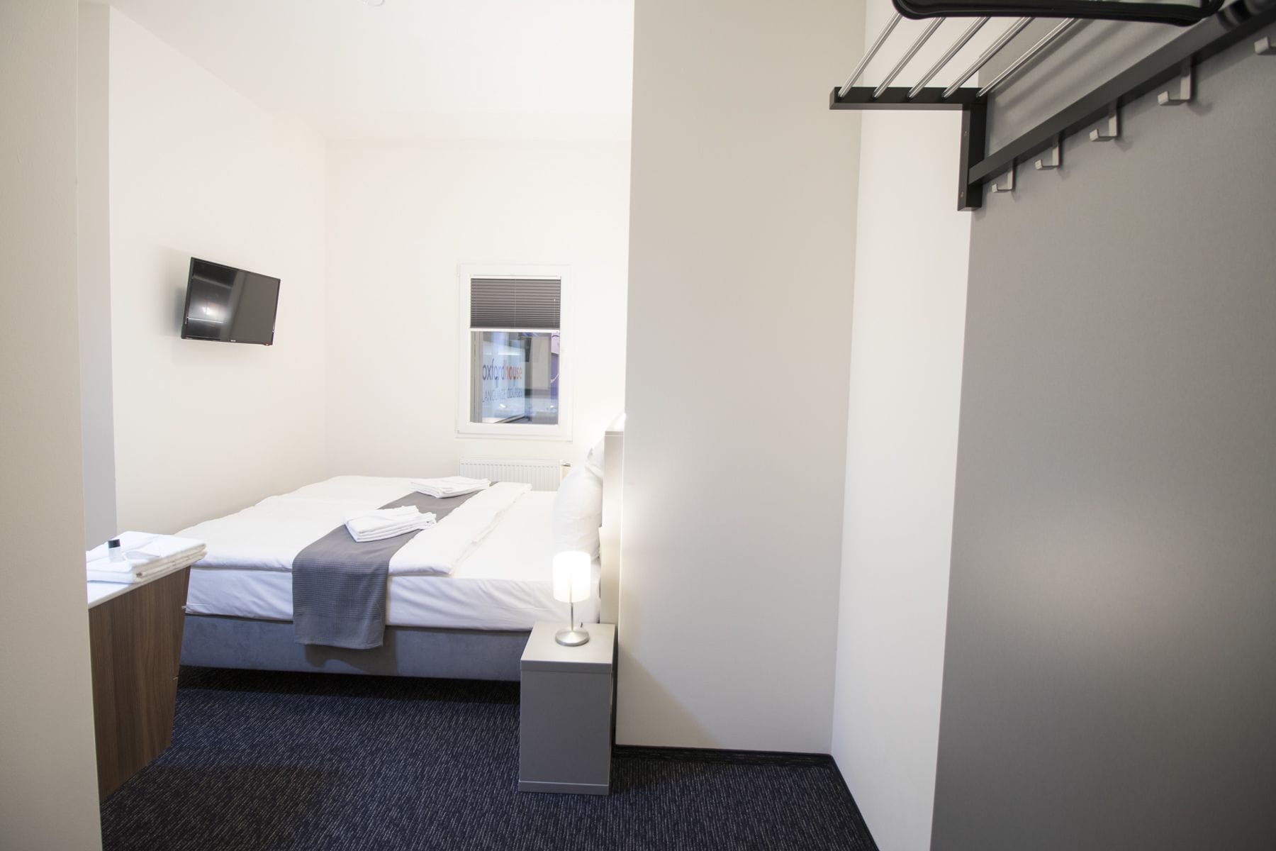 A hotel room with a bed, bedside table, lamp, and a window with blinds.