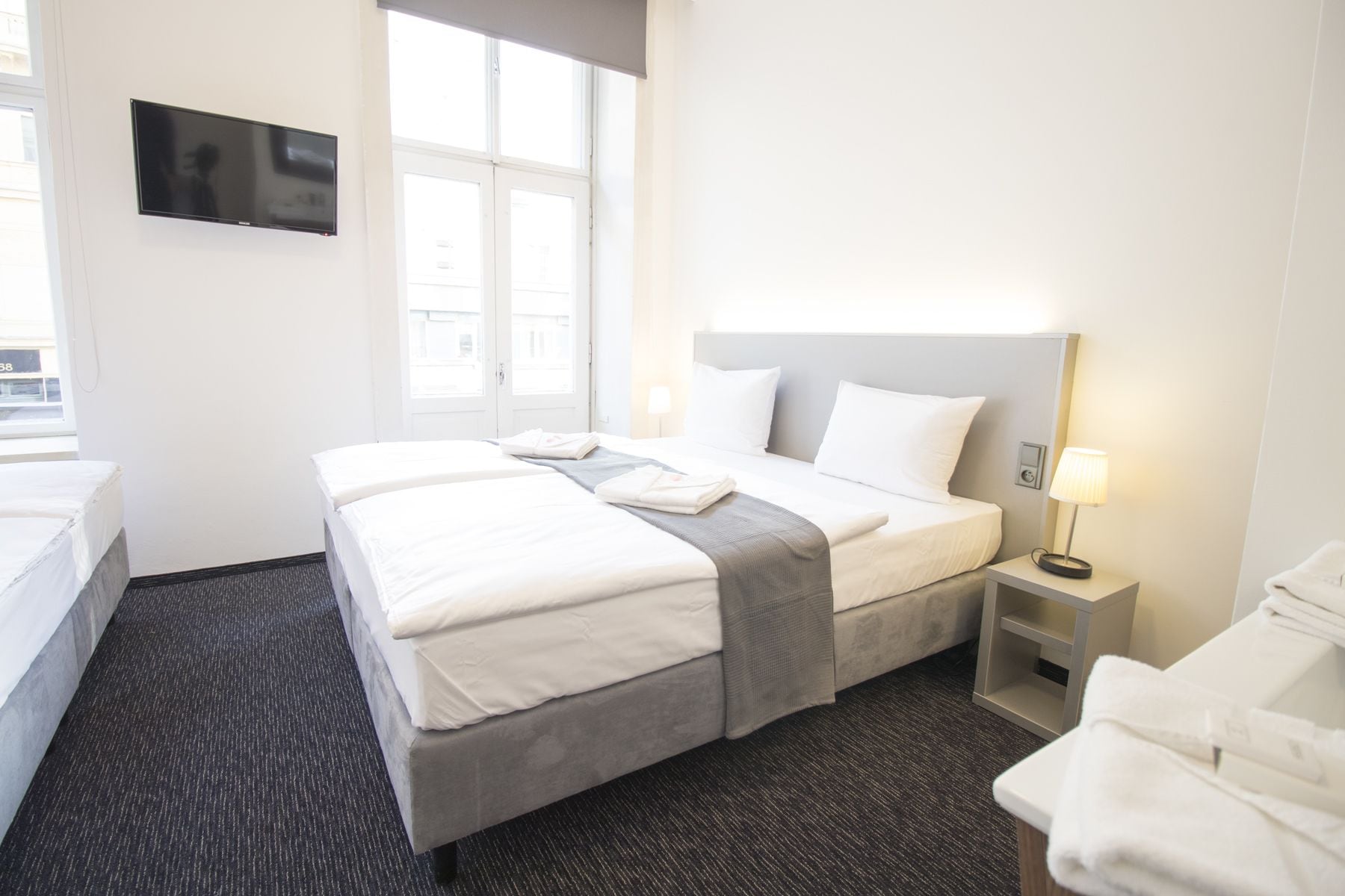A hotel room with a bed, a TV mounted on the wall, a window, a bedside table with a lamp, and a chair.