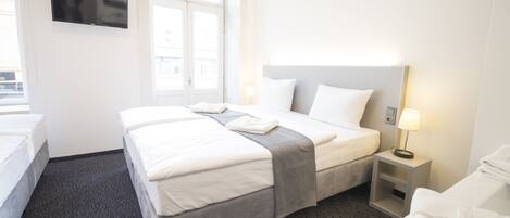 A hotel room with a bed, a TV mounted on the wall, a window, a bedside table with a lamp, and a chair.