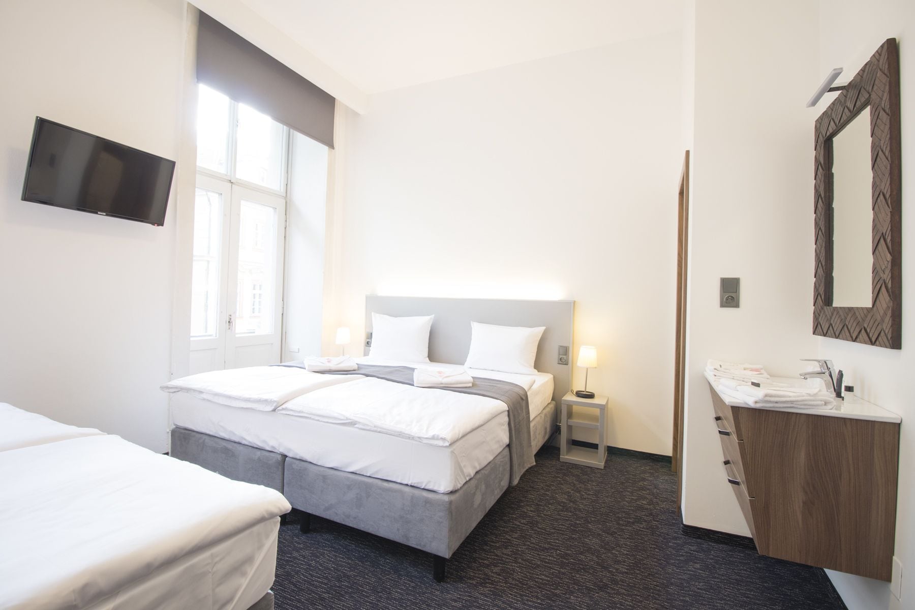 A hotel room with a bed, a bedside table with a lamp, a TV mounted on the wall, and a wooden cabinet with a mirror.