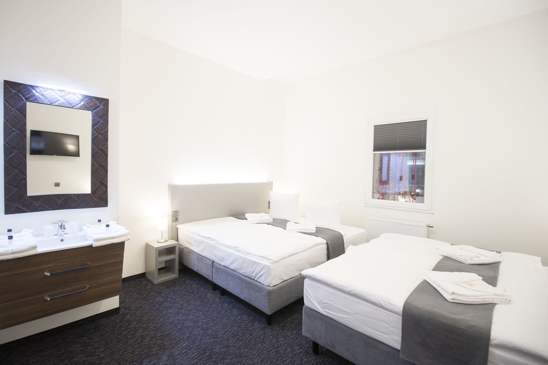 A hotel room with two beds, a large mirror, a TV, a bathroom with a sink and toiletries, and a window with blinds.