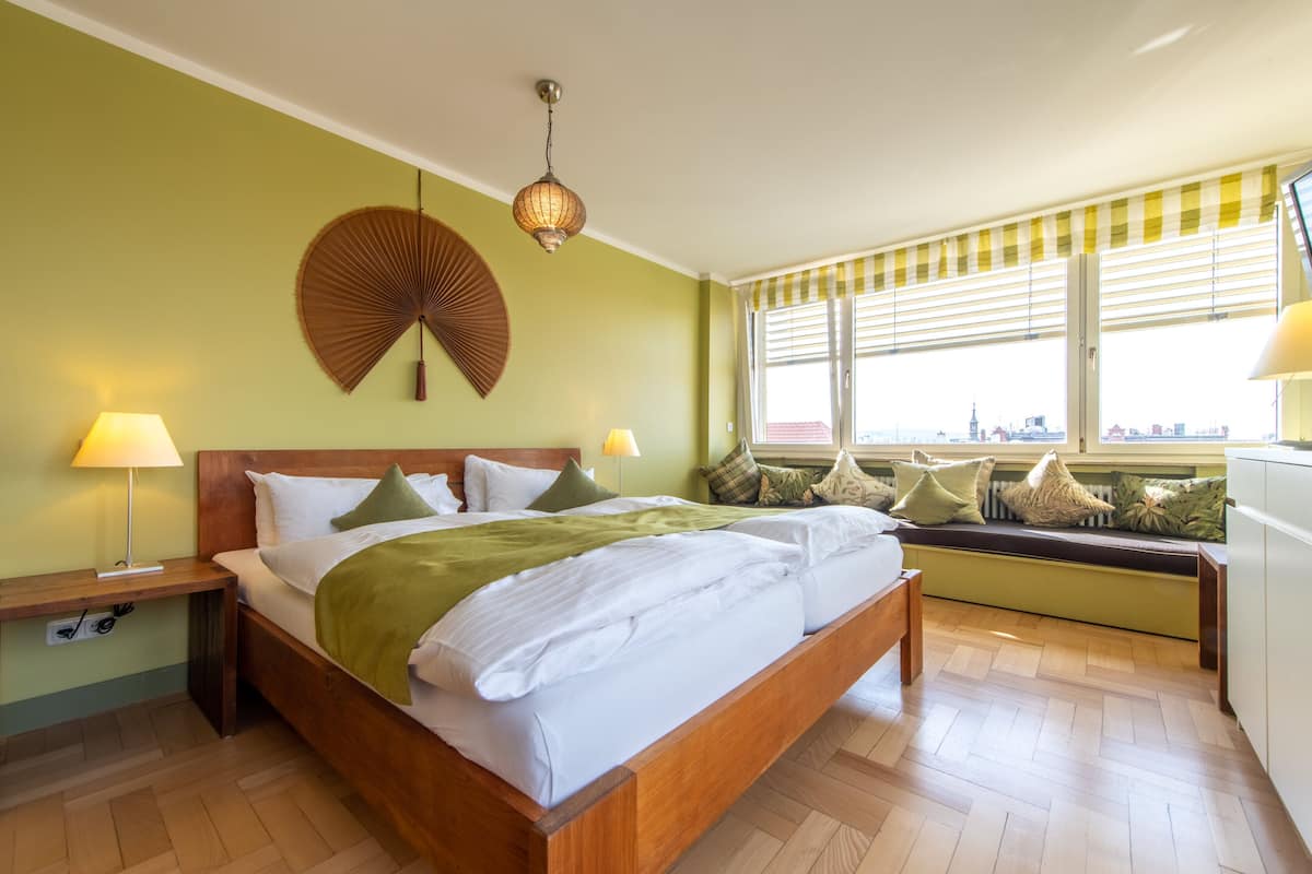 A bedroom with a wooden bed, green walls, a large window with blinds, and a city view.