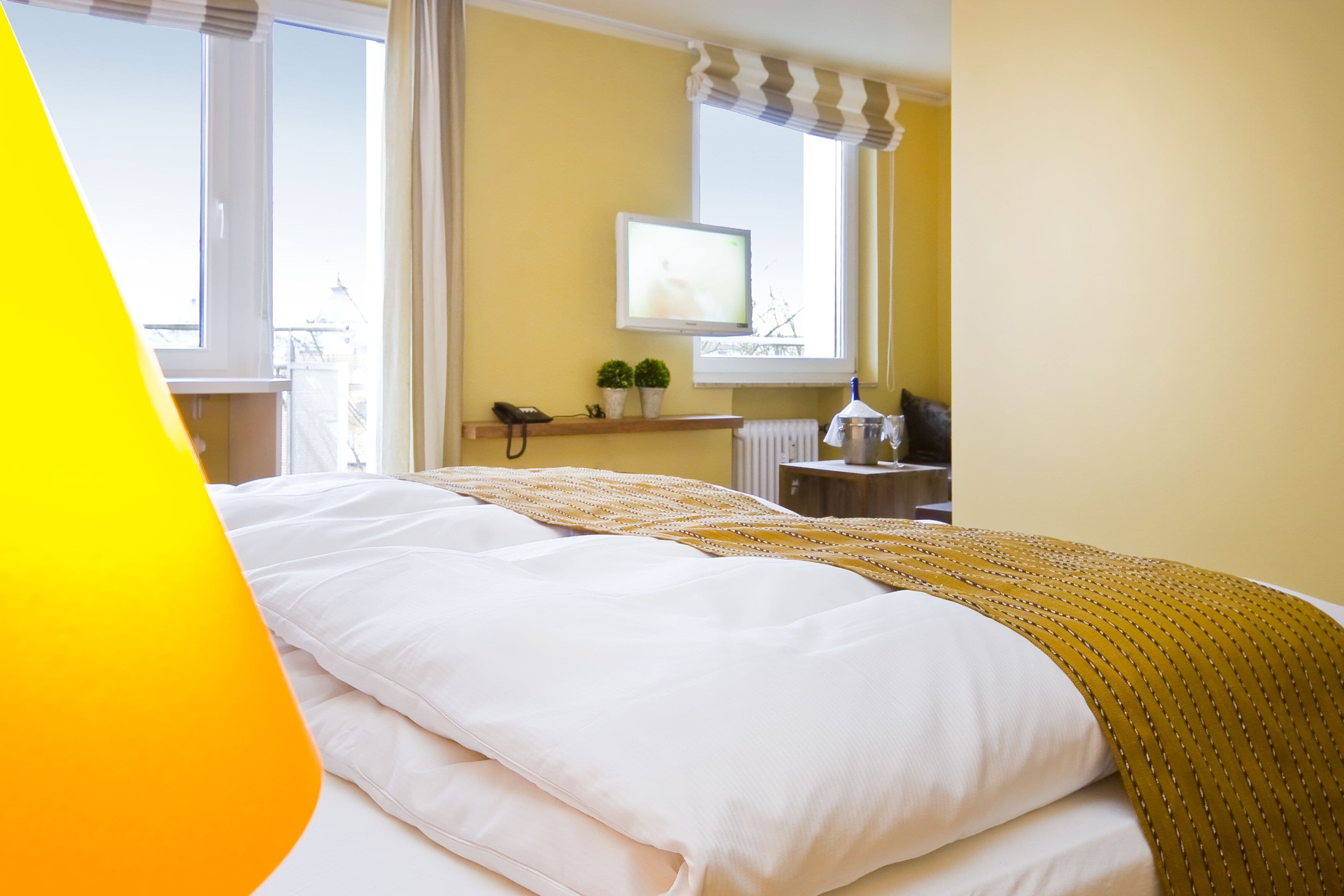 Een hotelkamer met een bed, een raam met gordijnen, een klein tafeltje met een telefoon en een plant, en een radiator.