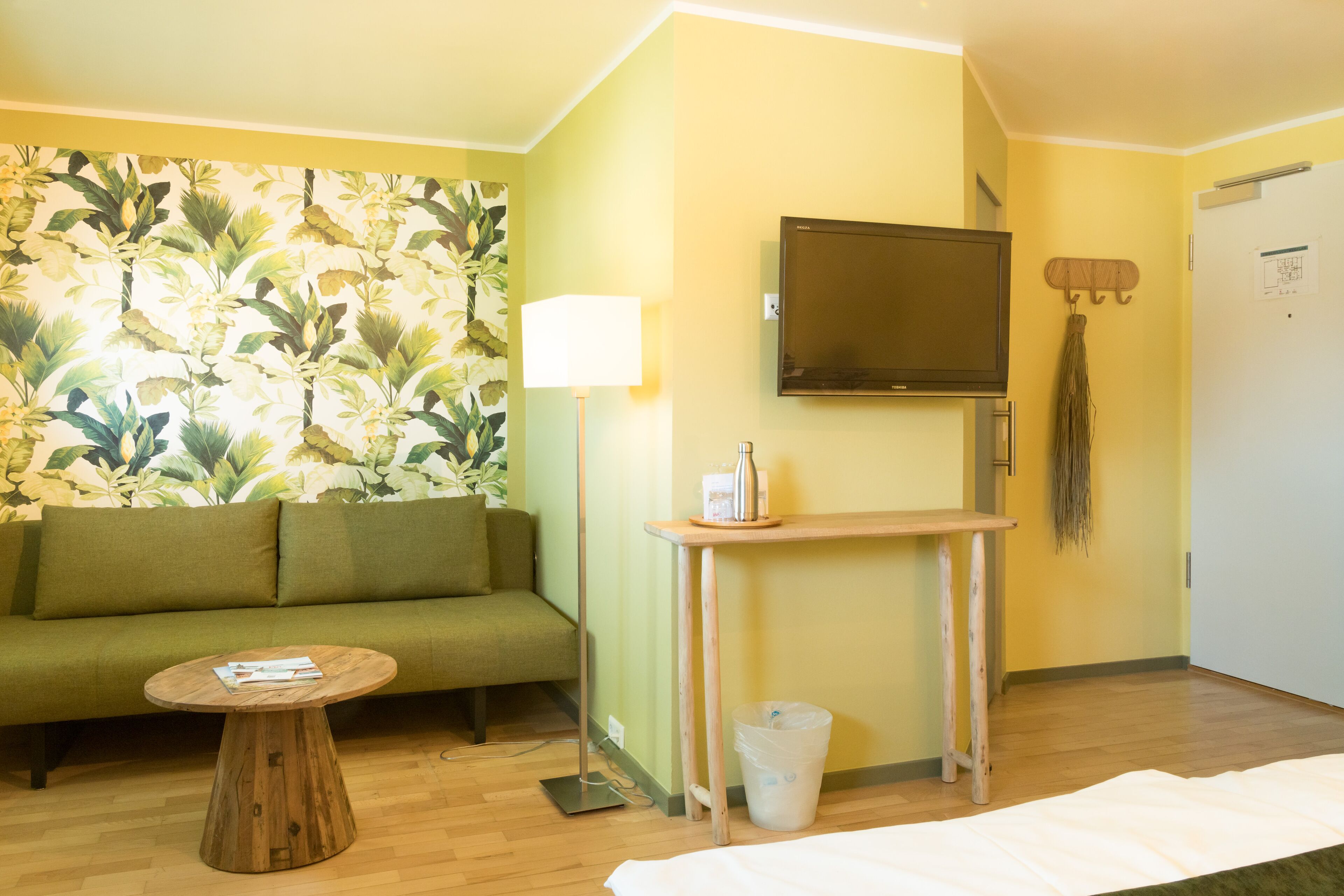 A room with a green sofa, a wooden coffee table, a TV mounted on the wall, a small table with a lamp, and a broom hanging on the wall.