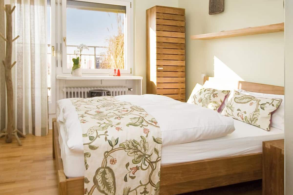 A bedroom with a wooden bed, a radiator, a window with curtains, and a shelf with decorative items.
