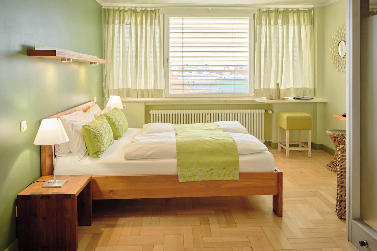 A bedroom with a wooden bed, bedside lamps, a window with curtains, a radiator, and a small stool.