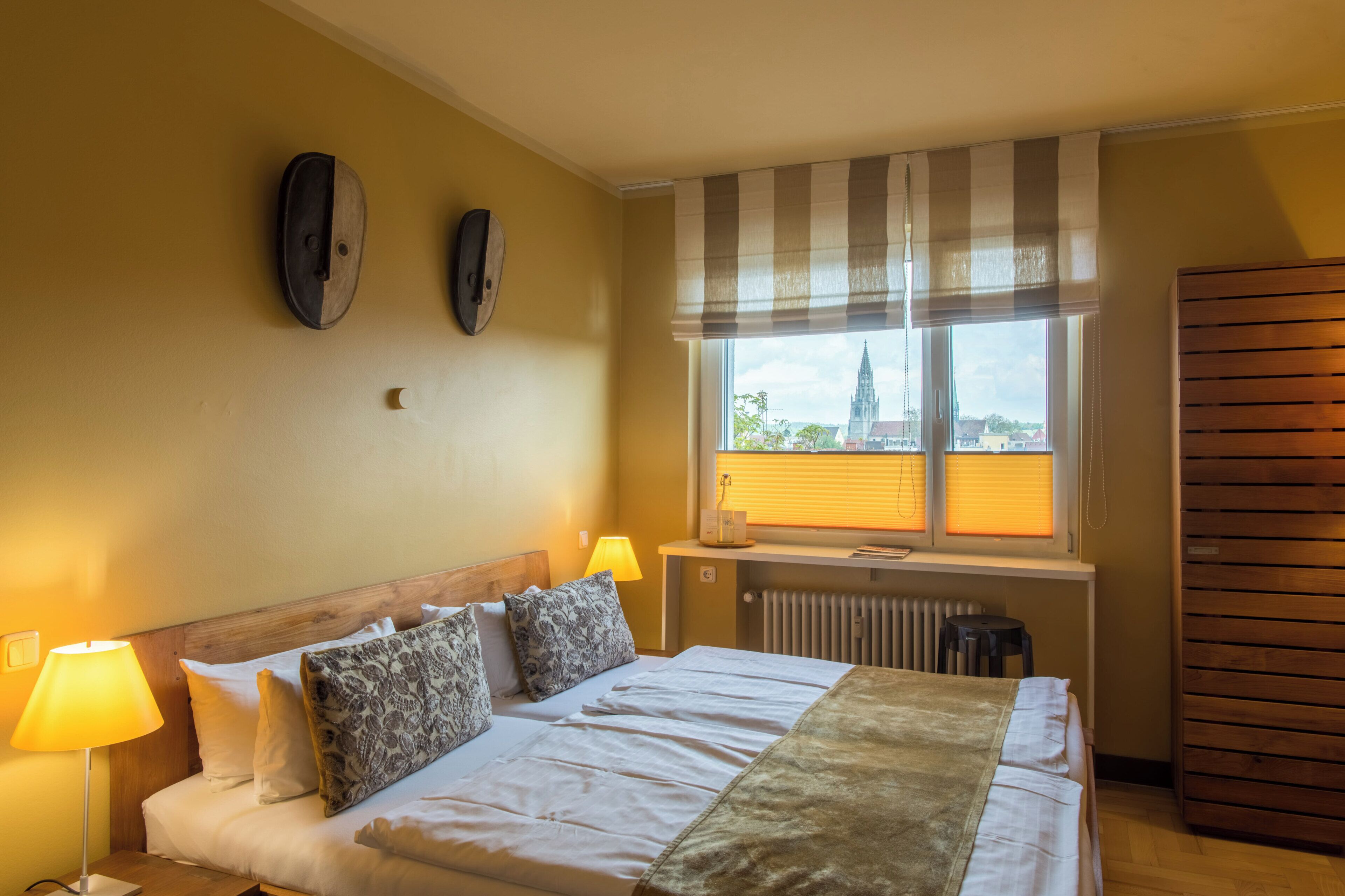 A hotel room with a bed, two bedside lamps, a window with blinds, a radiator, and a view of a distant building.