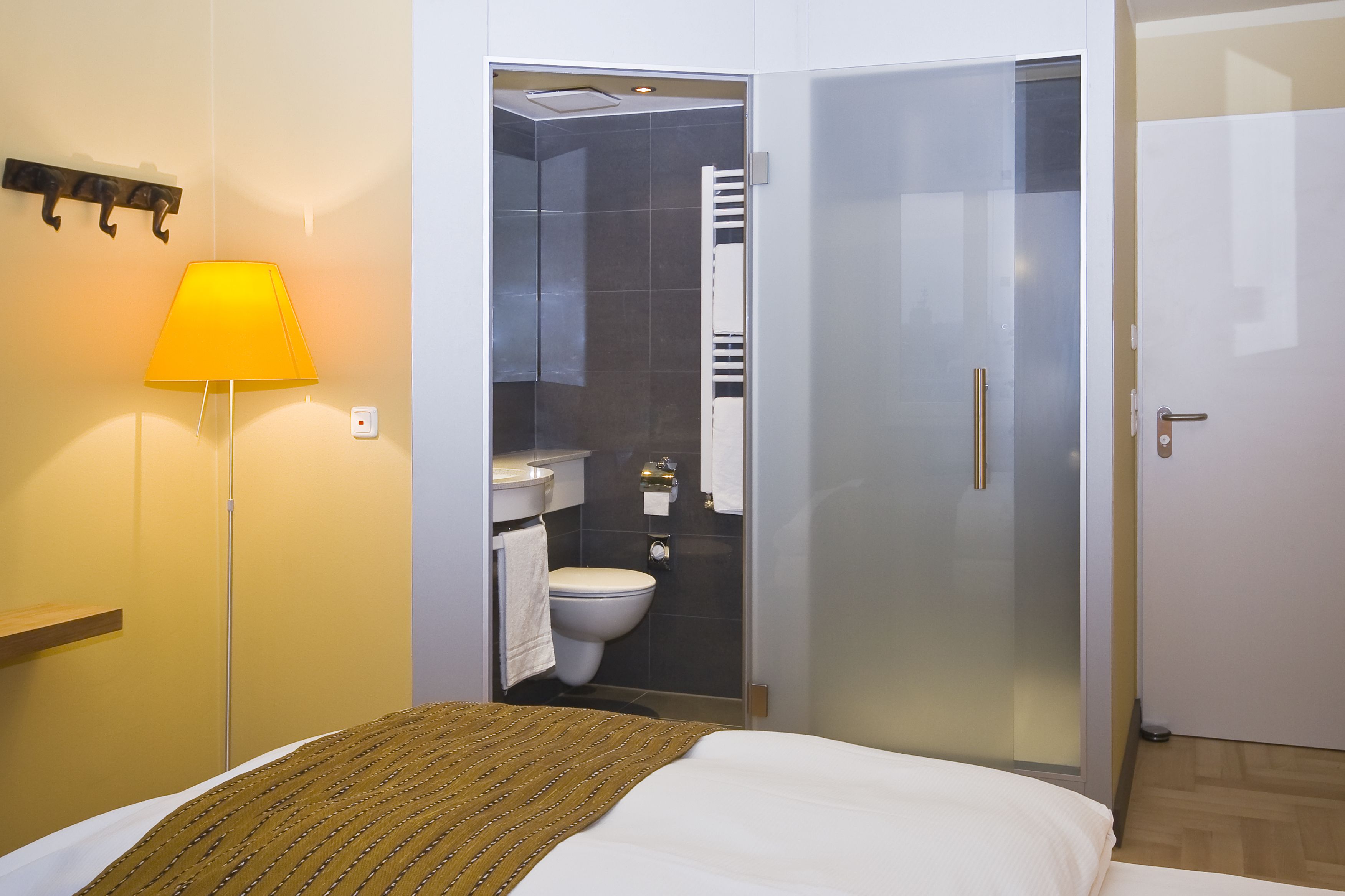 Ein Hotelzimmer mit Bett, Nachttischlampe, einer Duschkabine aus Glas und einer weißen Tür.
