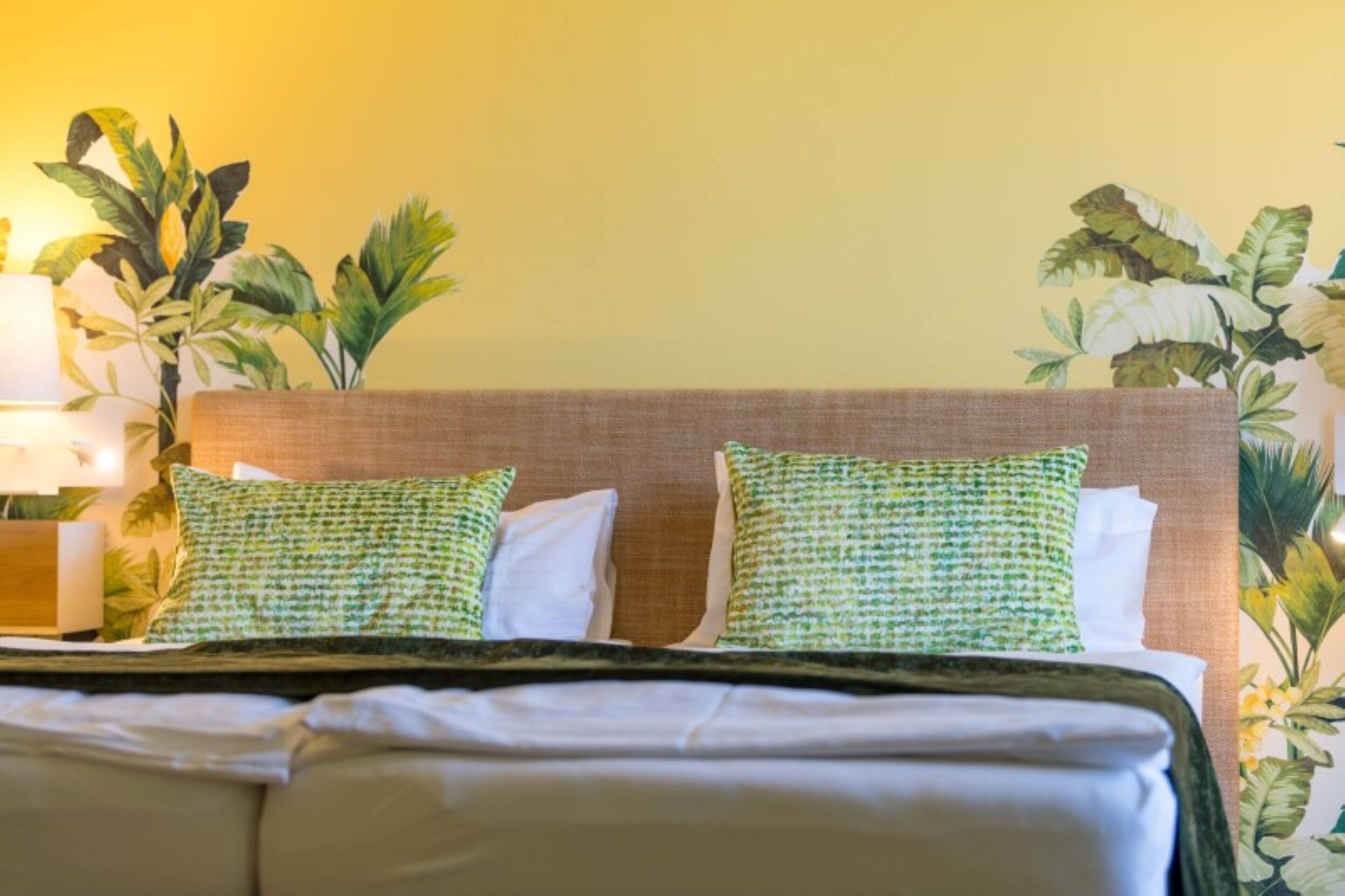 Habitación de hotel con cama, dos almohadas, mesita de noche con lámpara y una planta grande.
