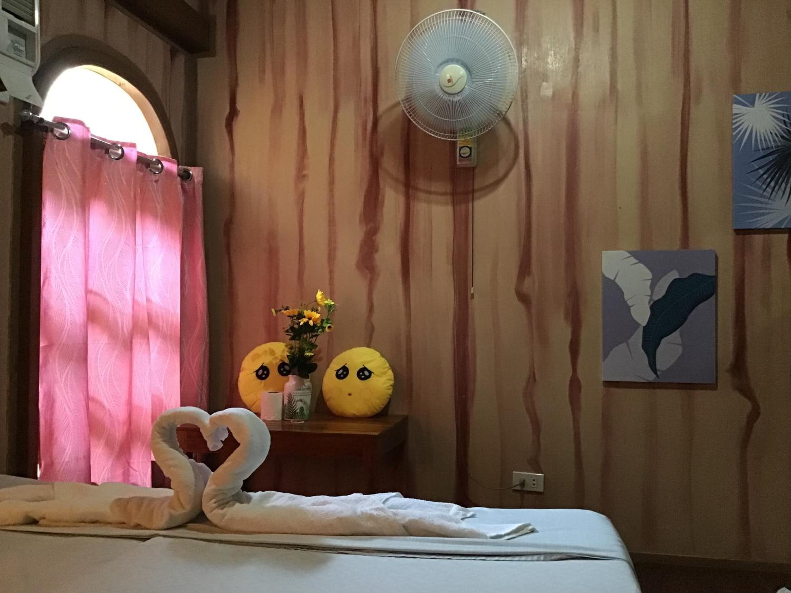 Habitación de hotel con cama, ventilador de pared, ventana con cortinas y una mesita con un jarrón de flores y dos almohadones decorativos.