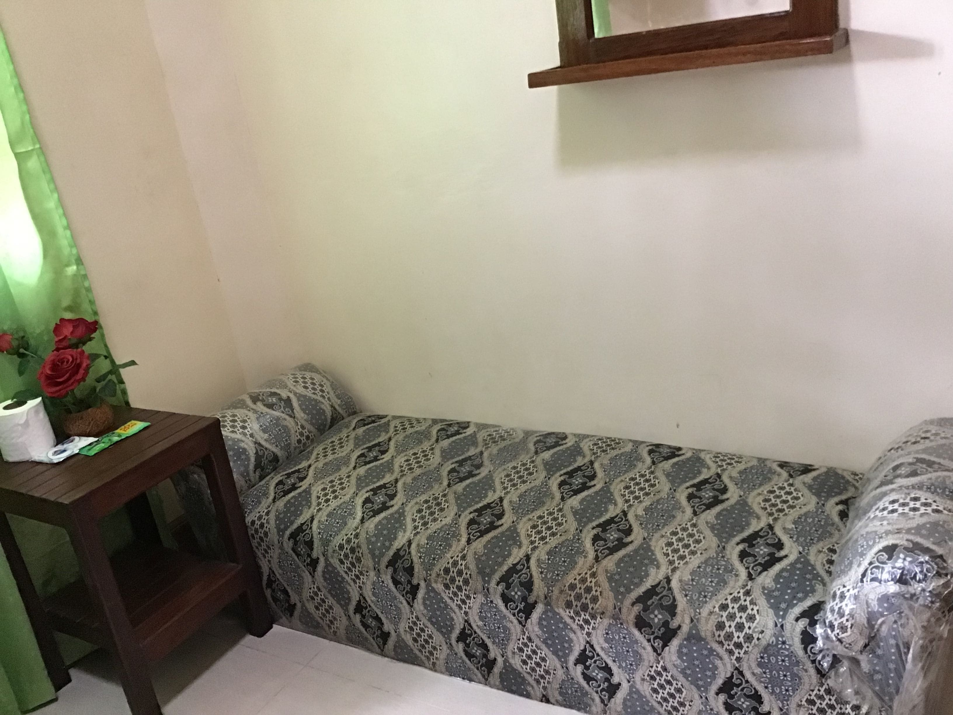 A corner room with a patterned sofa, a small wooden table with a tissue box and a packet, and a green curtain with a rose decoration.