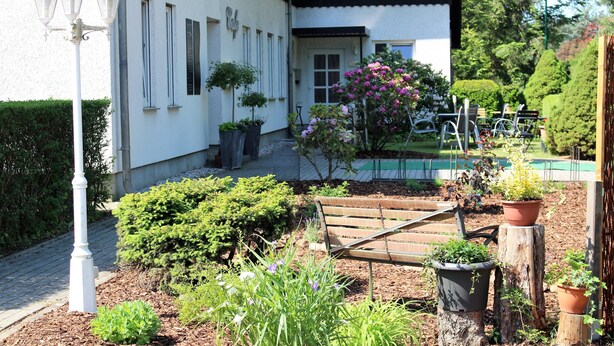 A garden with a wooden bench, potted plants, and a white building with a red roof. - Pension & Restaurant Nordstern (Cottbus)