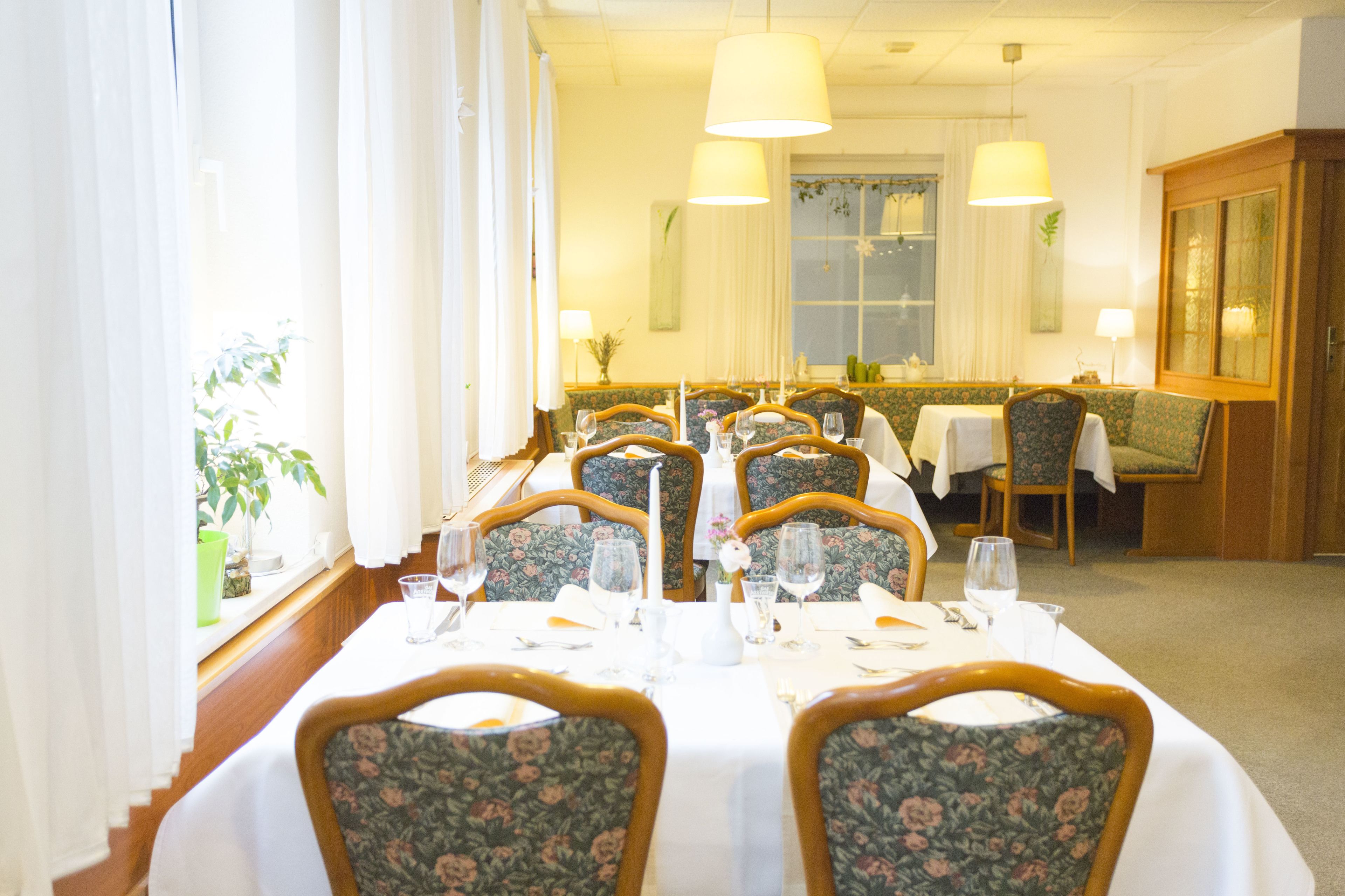 A dining area with multiple tables set for service, chairs with floral upholstery, and a large window with curtains.